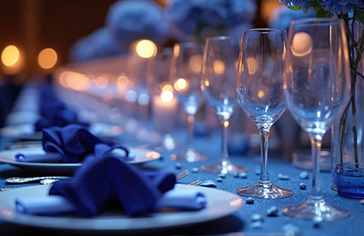 Elegant gala table setting with blue accents. White plates, napkins, silverware, wine glasses, lights.