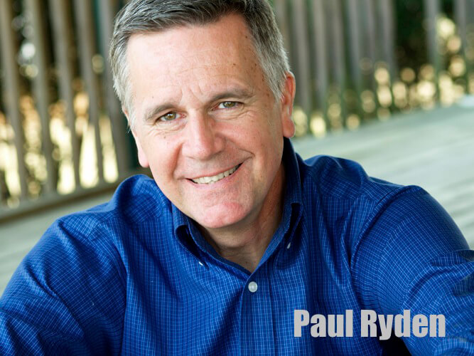 Image of Paul Ryden sitting and smiling at the camera wearing a blue button up shirt.