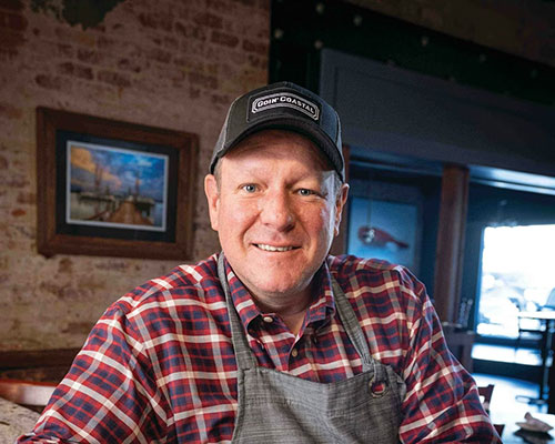 Chef Zachary Kell wearing a black trucker style hat, and a chef apron with a brick restaurant wall in the background
