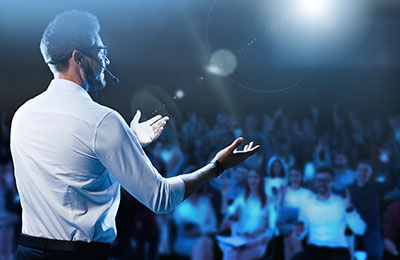 A man with a headset on with arms out standing on a stage speaking to an audience in the background